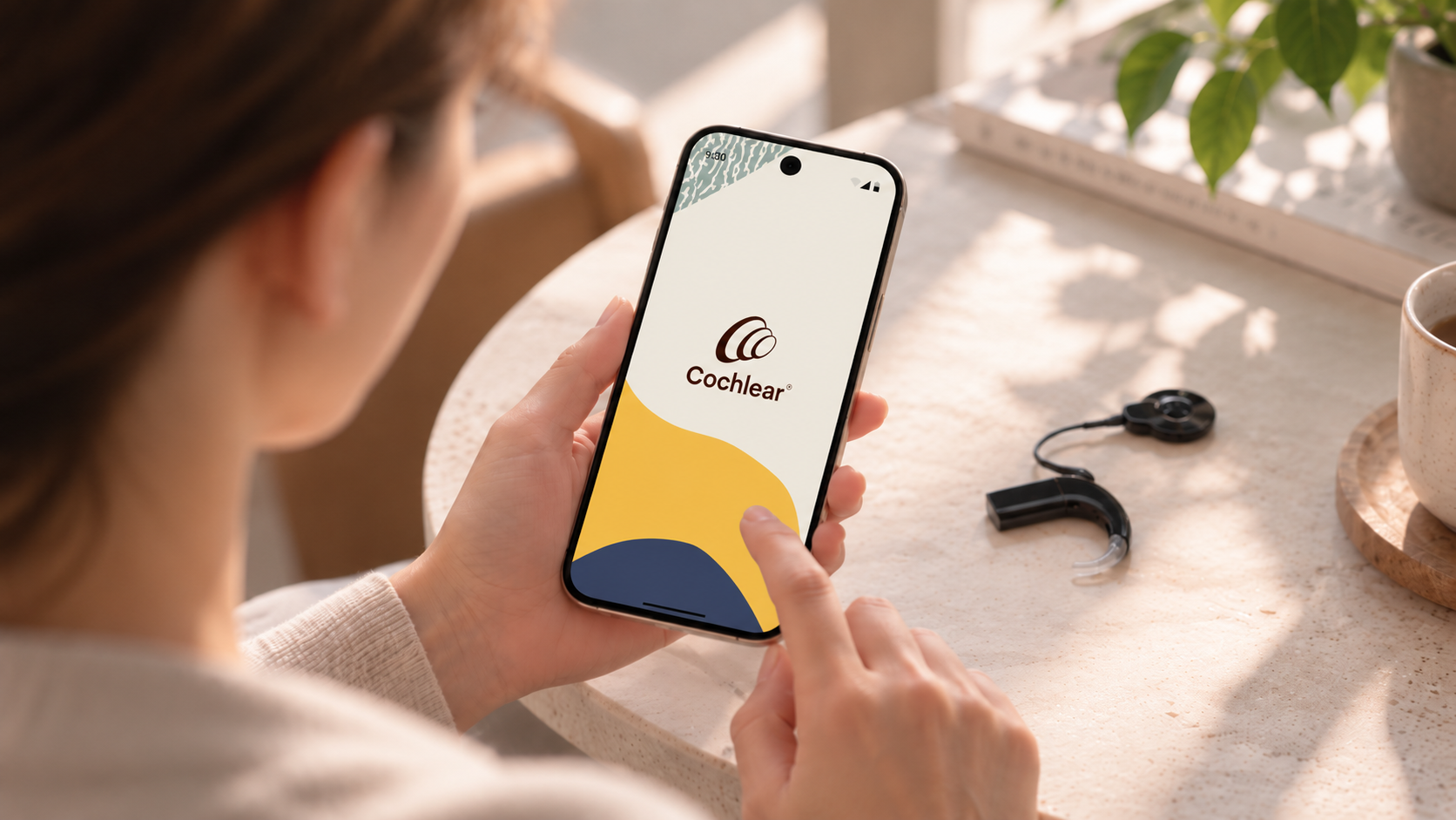 A person opening the Nucleus Smart App on their phone, with a Cochlear hearing device on the table